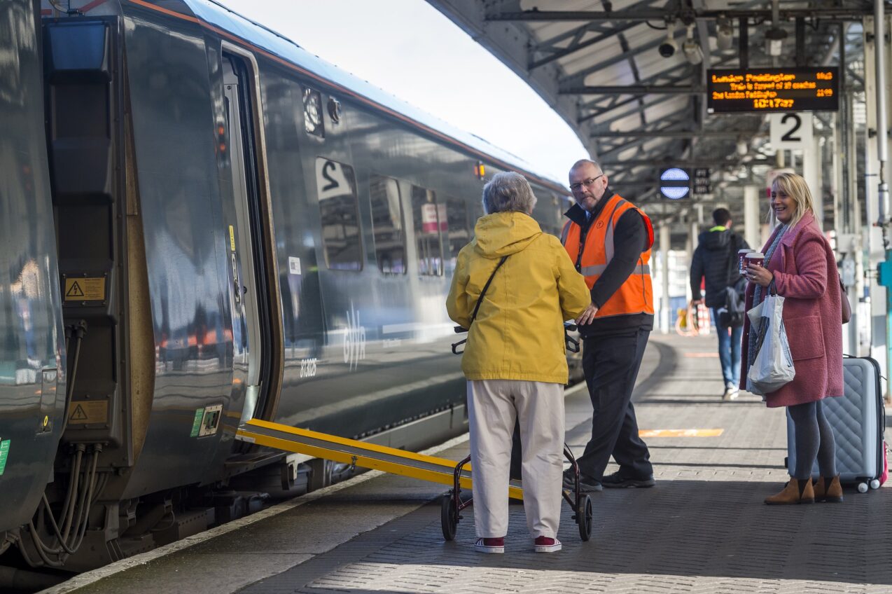 Staff member assisting passenger with mobility aid on to train with ramp deployed at a train station