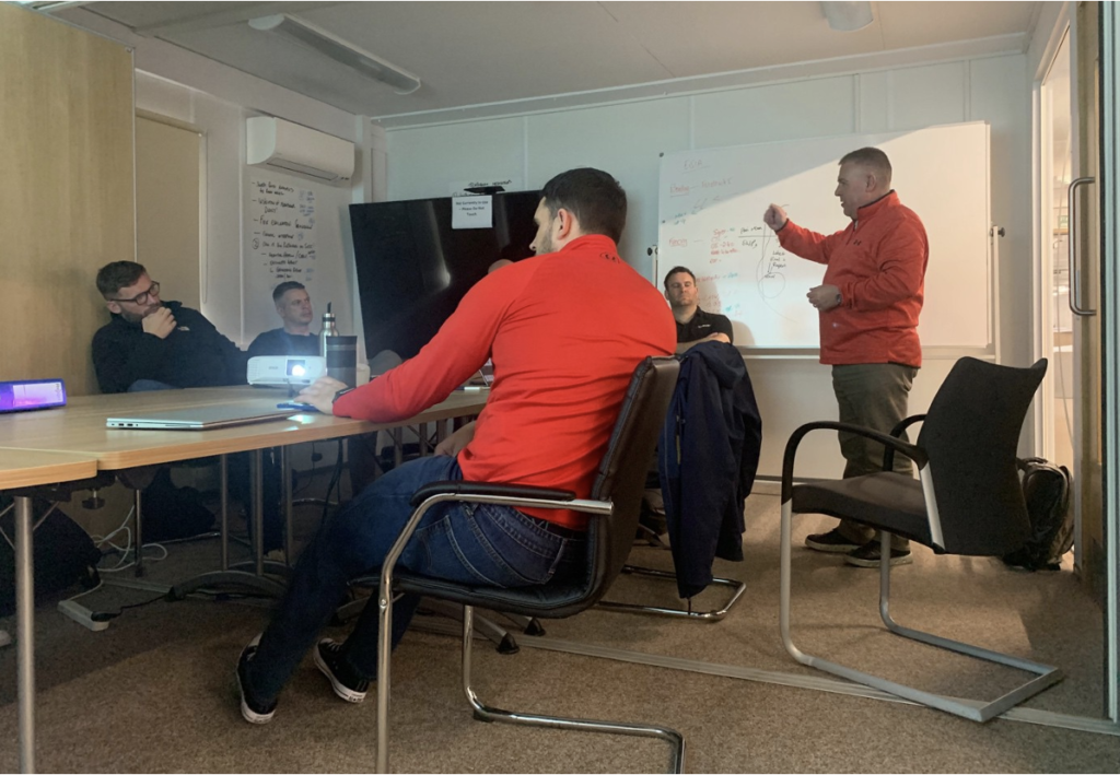 A group of people in a classroom setting listening to a man stood instructing them all by a whiteboard