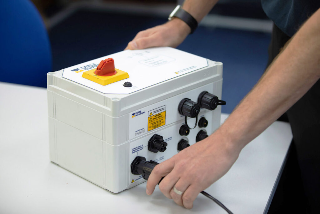 A person is plugging a power cable into the cable guardian. It's a white box with many ports on the front and a red switch on top