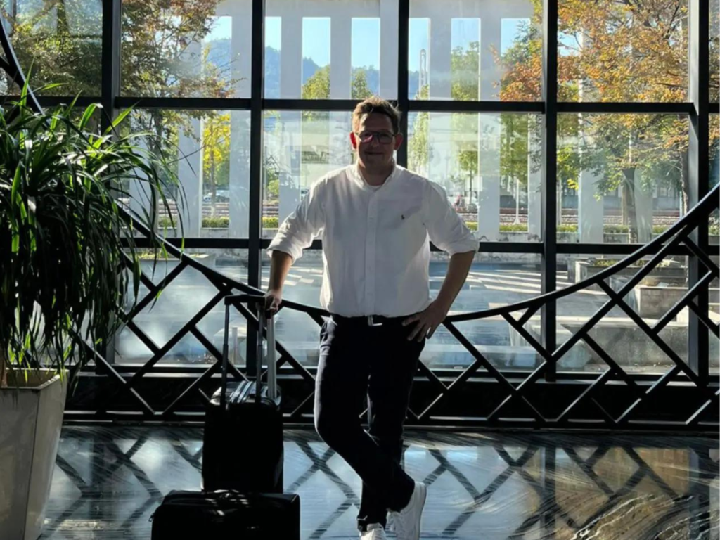 A man in a white shirt, black trousers and white sneakers standing with his luggage in front of a large window