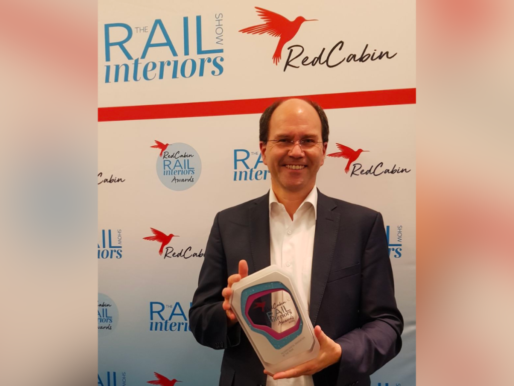 A man in a suit smiles and holds a grey, blue and purple award. The background is covered in red cabin rail interior show logos