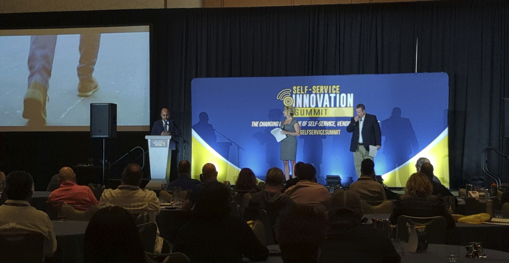 There are three people stood on a stage in front of a seated audience. The background is dark blue with yellow and white text reading "Self-Service Innovation Summit"