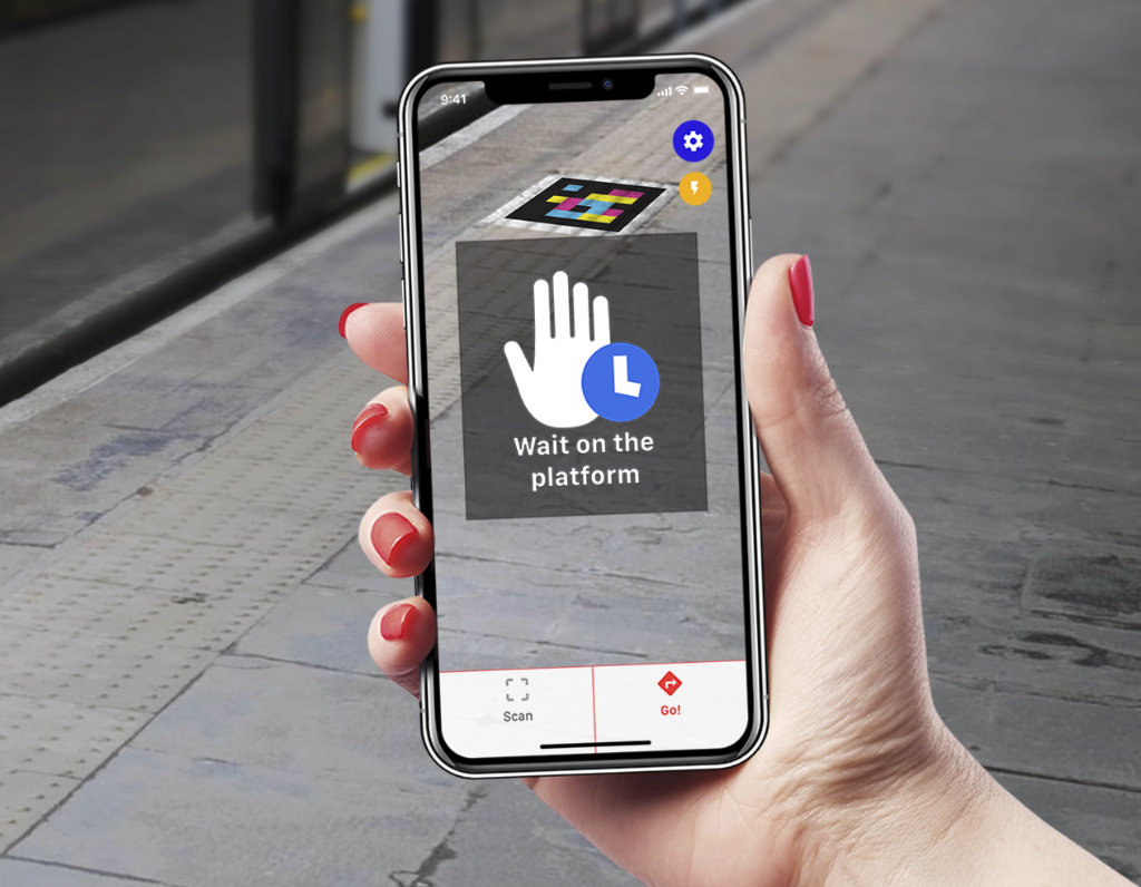 A hand with red nail polish holds a phone, pointed at a train track. On the phone we can see a colourful QR code on the platform floor. There's an overlay which reads "wait on the platform"