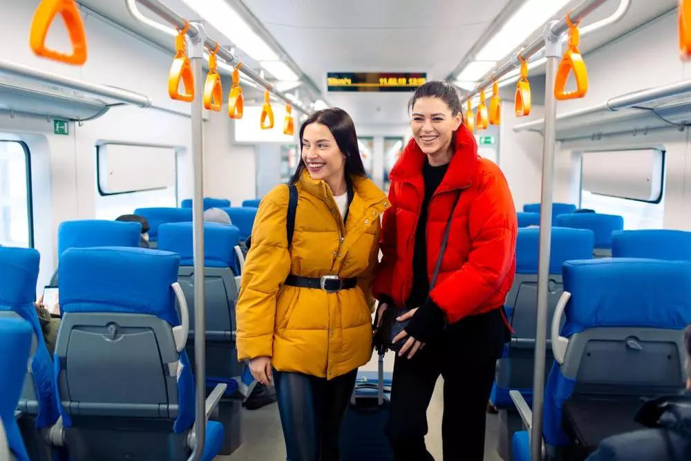 Two smiling women standing in a train isle with their luggage. One woman has her long brown hair down and is wearing a yellow coat. The other woman has brown hair in a ponytail and is wearing a red coat