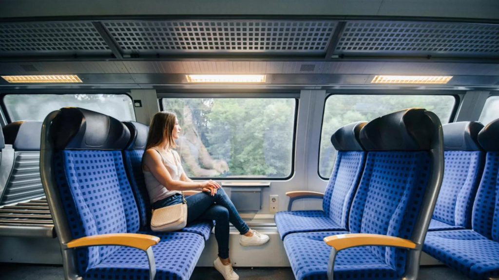 A woman sits on blue train seats looking out the window. There are heavily packed trees in the background