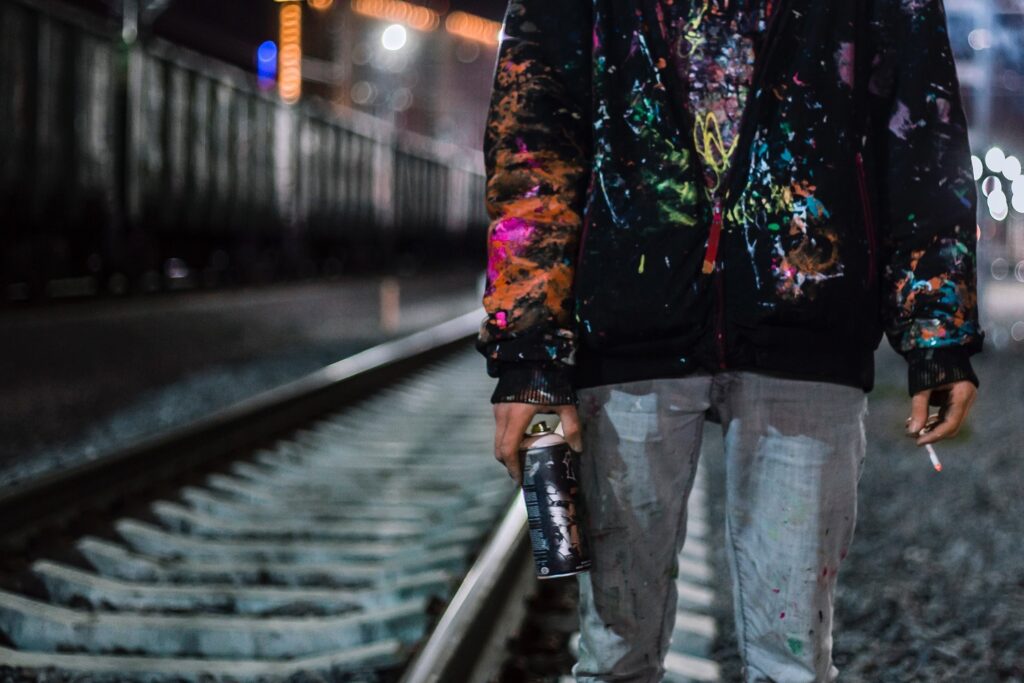 In the front right of the image is a person (from chest to knees) in jeans and a black hoodie which is covered in colourful paint splotches. The person is holding a spray paint can in one hand and a lit cigarette in the other. Behind them to the left is a rail track