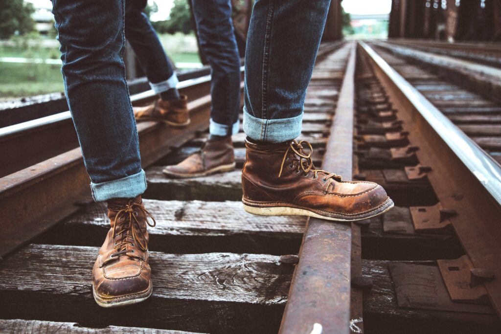 Two people in brown boots and cuffed jeans stand on a train track. We can only see their ankles and feet.