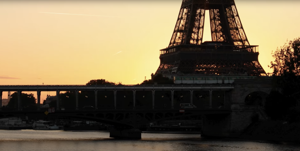 The eiffel tower is in silouette with an orange sunset sky behind it. There is a train travelling on a track in the foreground