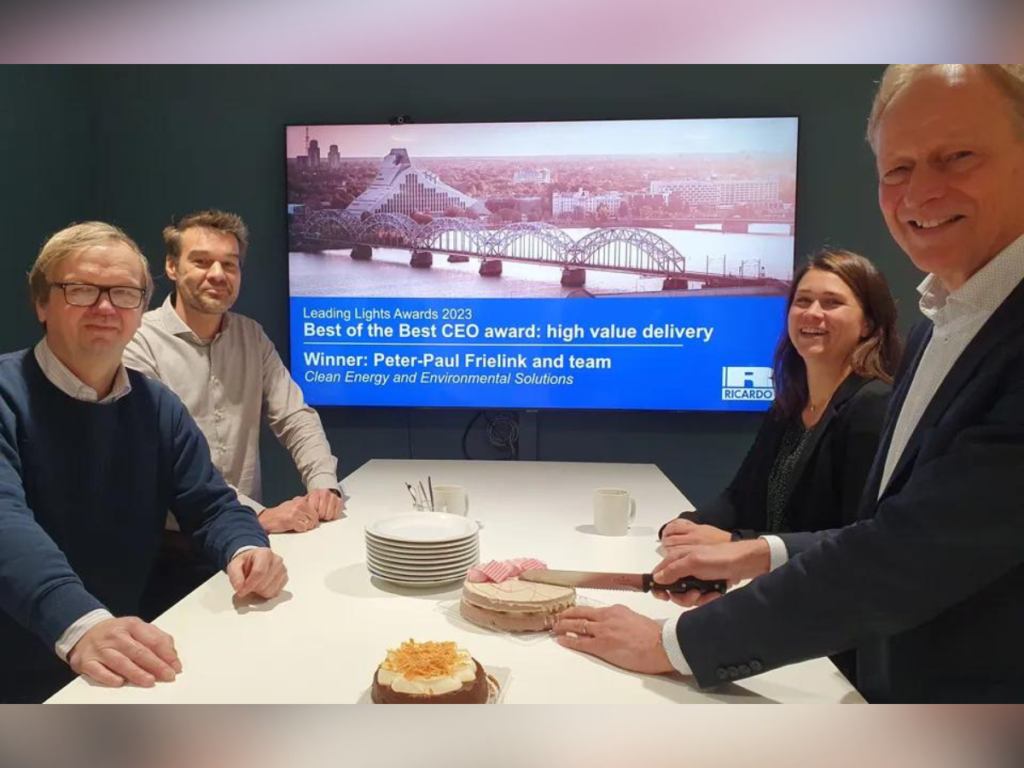 Four people sit around a white table. There are two cakes, some plates and mugs. In the background there is a monitor displaying a bridge and text reading "Leading Lights Awards 2023: Best of the Best CEO Award: High Value Delivery. Winner: Peter-Paul Frielink and Team