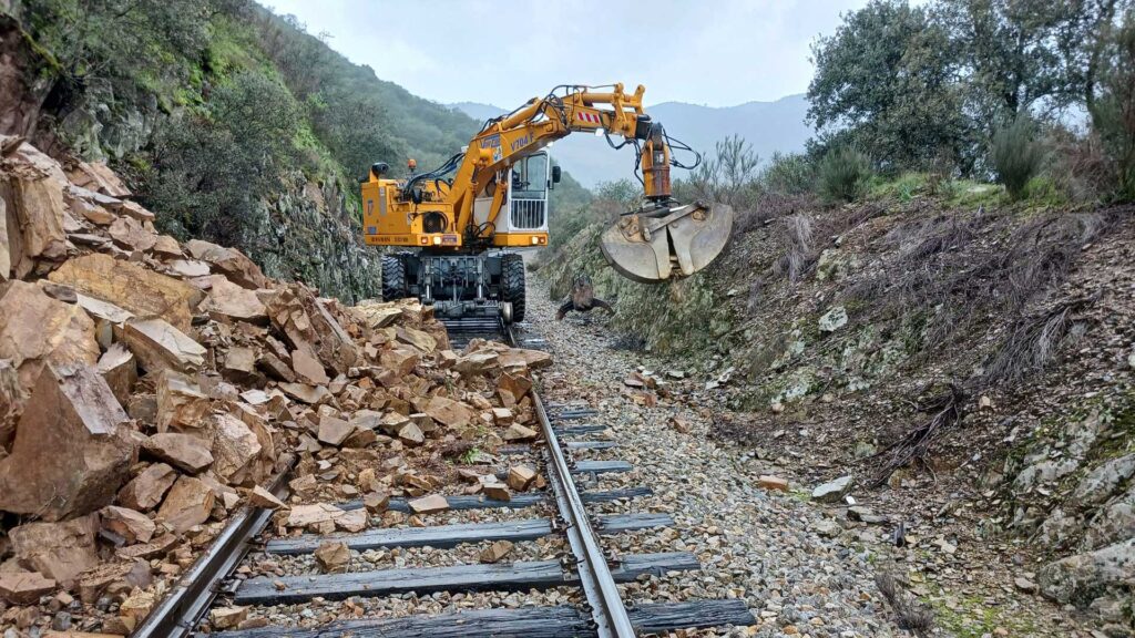 A landslide has covered a railway track in large rocks. There is a crane on the tracks removing the rocks