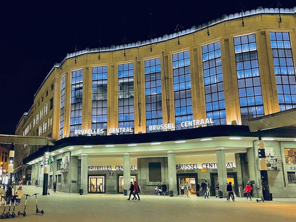 Brussels Central Station at night. The building is a light beige, there are rows of blue windows on the top half and white columns near the entrance