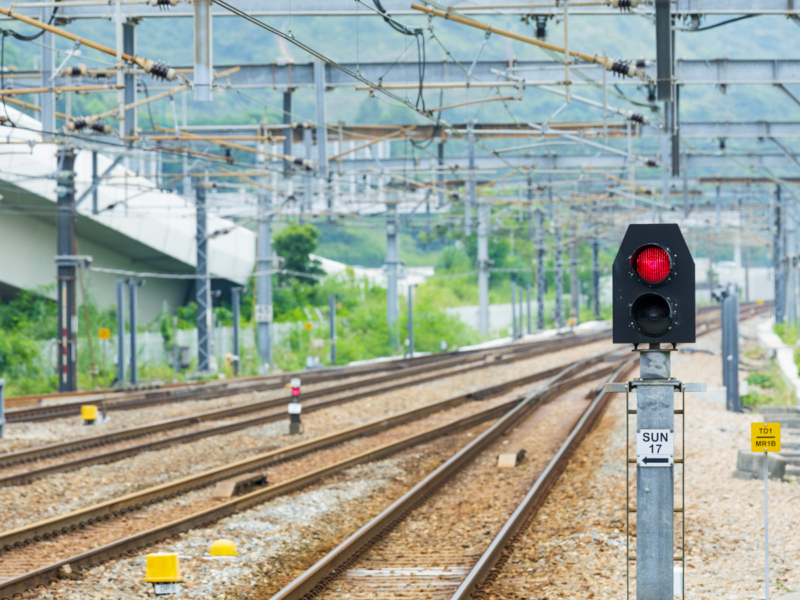 A railway signal displaying a red light next to multiple railway tracks, with overhead power lines and green hills in the background.
