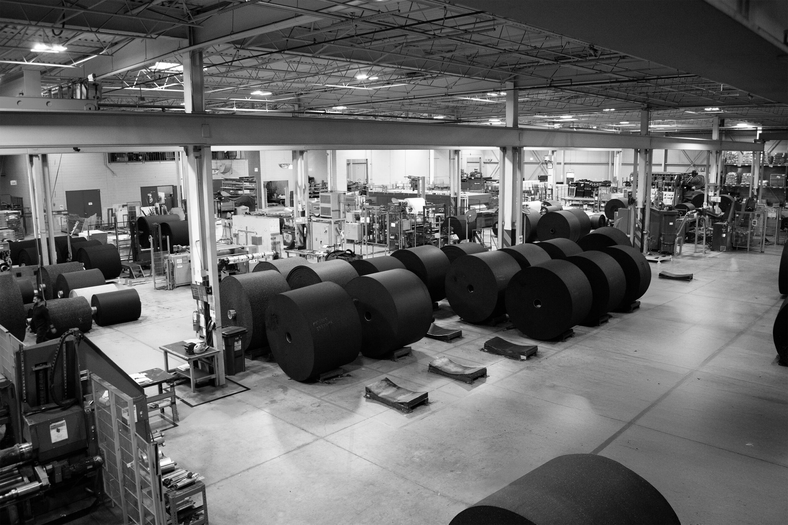 A black-and-white photo of a large industrial manufacturing facility, showing numerous massive rolls of rubber material arranged on the factory floor.