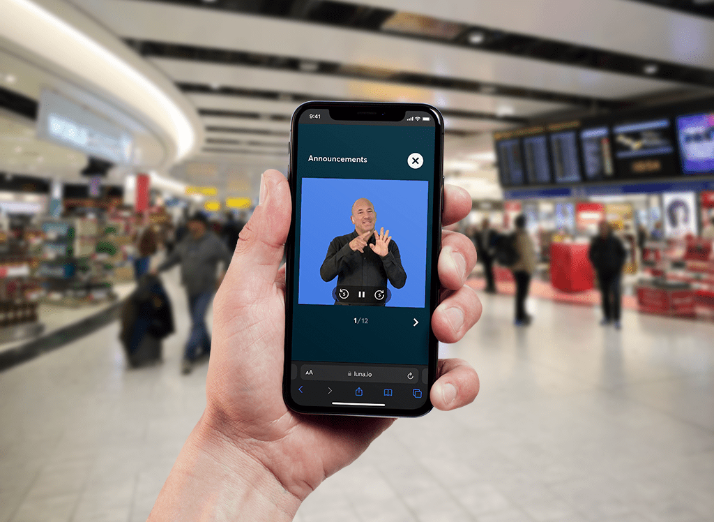 A person holding a smartphone in an airport terminal, displaying a sign language interpreter on the screen for airport announcements, with a busy terminal and flight information boards in the background.