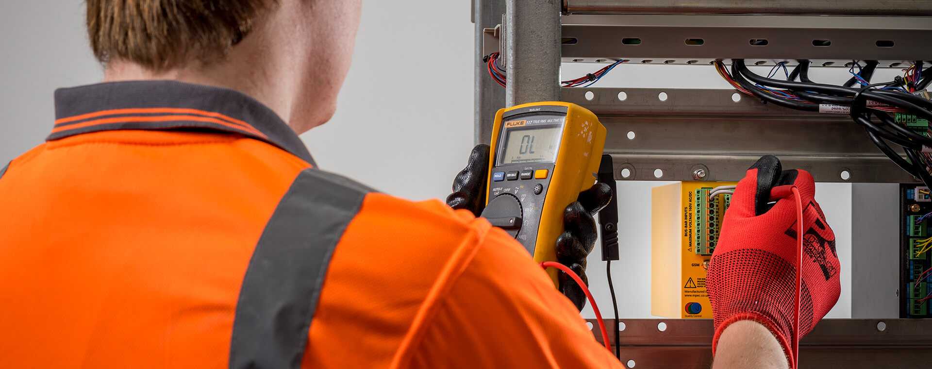 Technician in orange safety gear using a digital multimeter to test electrical connections inside an industrial control panel.
