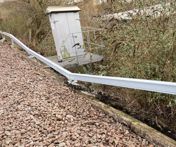 Damaged railway safety barrier beside a track, with nearby vegetation and residential buildings in the background.