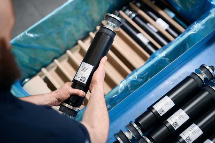 A person wearing a dark blue shirt and gloves is shown from over the shoulder, holding a black cylindrical suspension damper with a white label, with a blue bin full of similar dampers in the foreground and background.