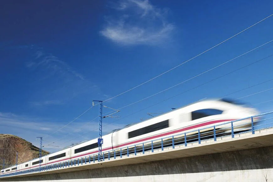 A long, sleek white high-speed train with red stripes is captured in motion blur as it speeds across a concrete bridge under a bright blue sky with wispy clouds.