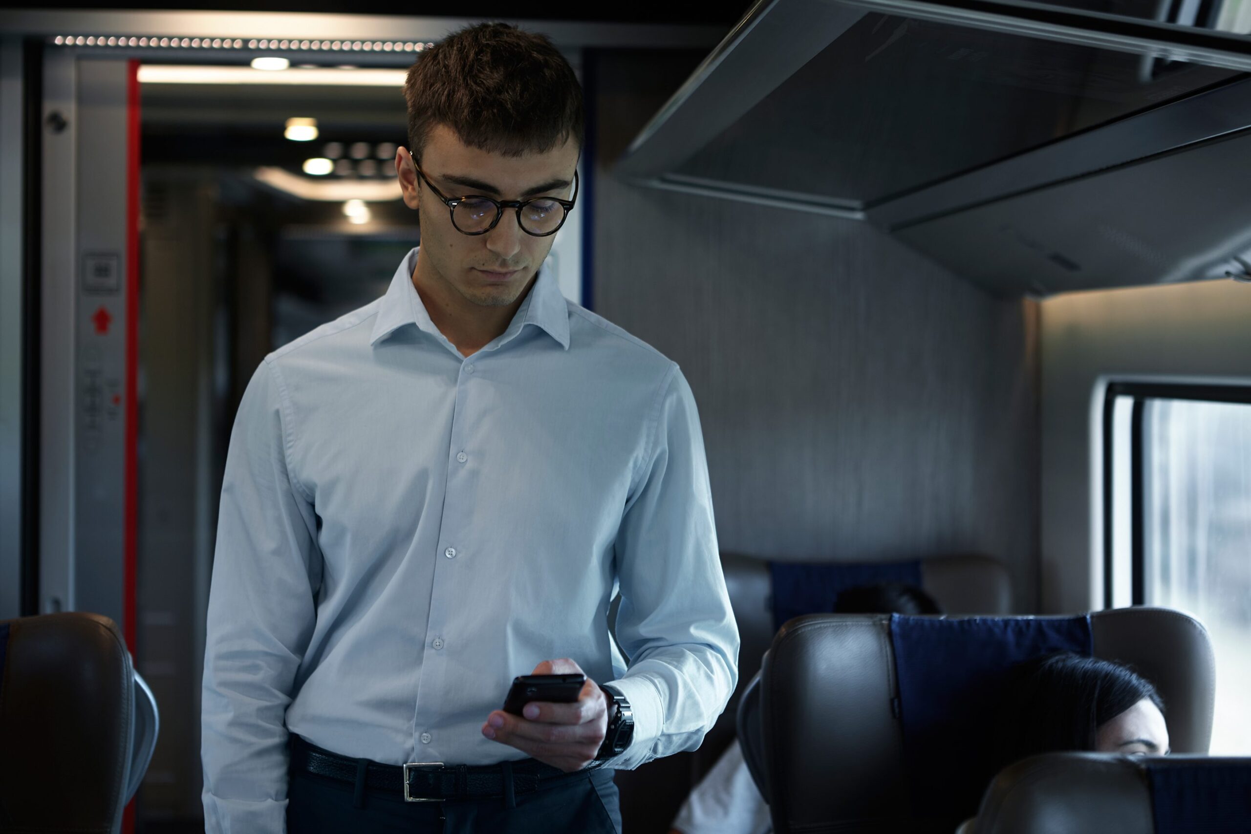 A young man with short dark hair and glasses stands in what appears to be a train car, looking down at his phone while wearing a light blue button-up shirt and dark pants.
