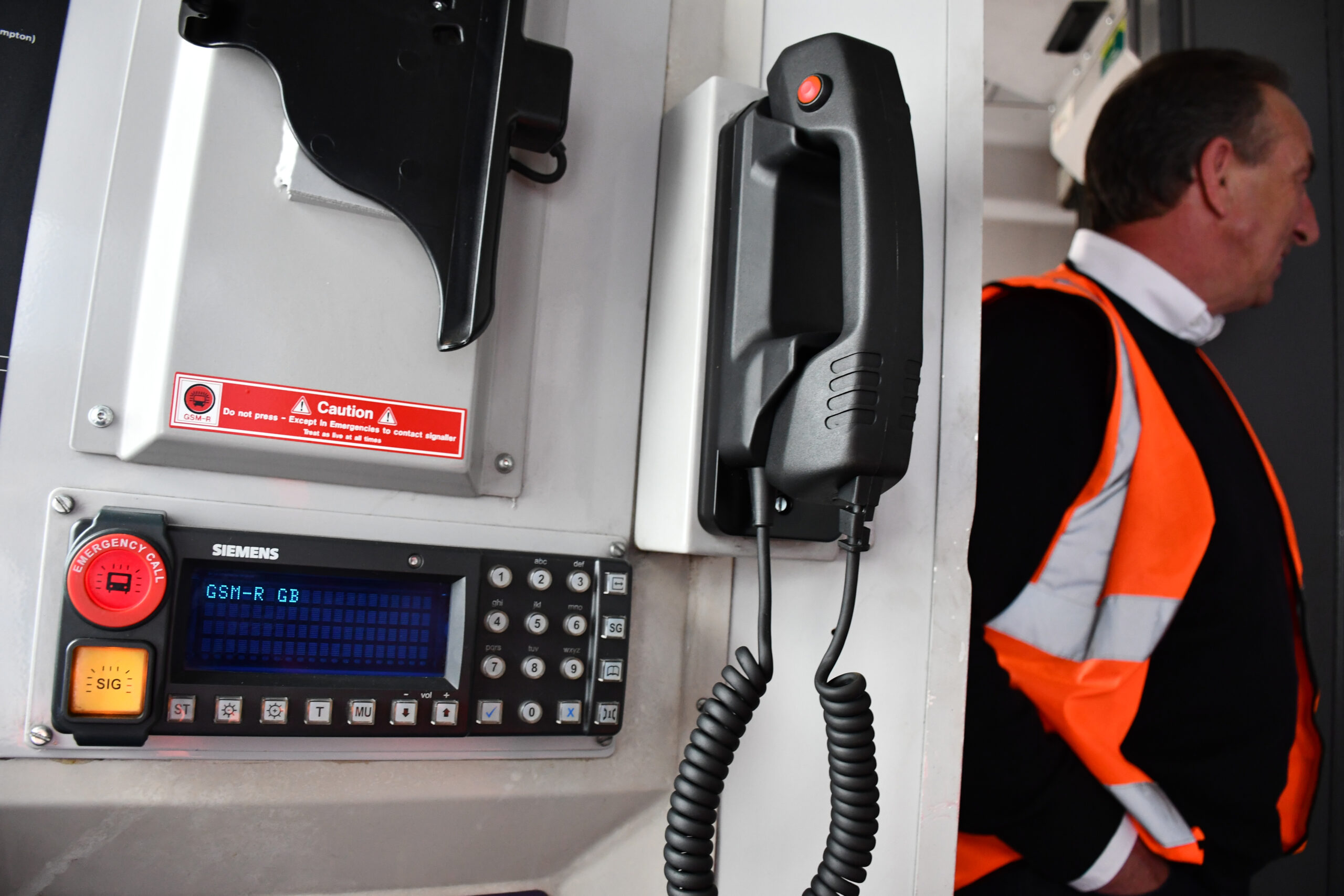 A close-up shot of a Siemens GSM-R GB communication system mounted on a wall, featuring a black handset with a coiled cord, an orange emergency call button, and a digital display, with a person in an orange high-visibility vest partially visible on the right.