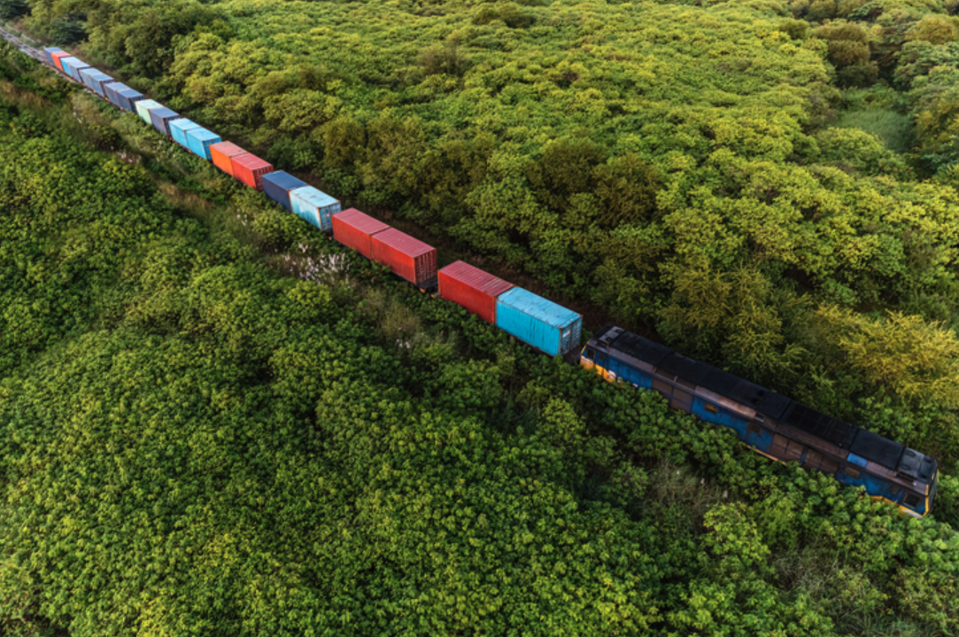 An aerial view shows a long freight train with various colored cargo containers traversing a track through a dense, lush green forest.