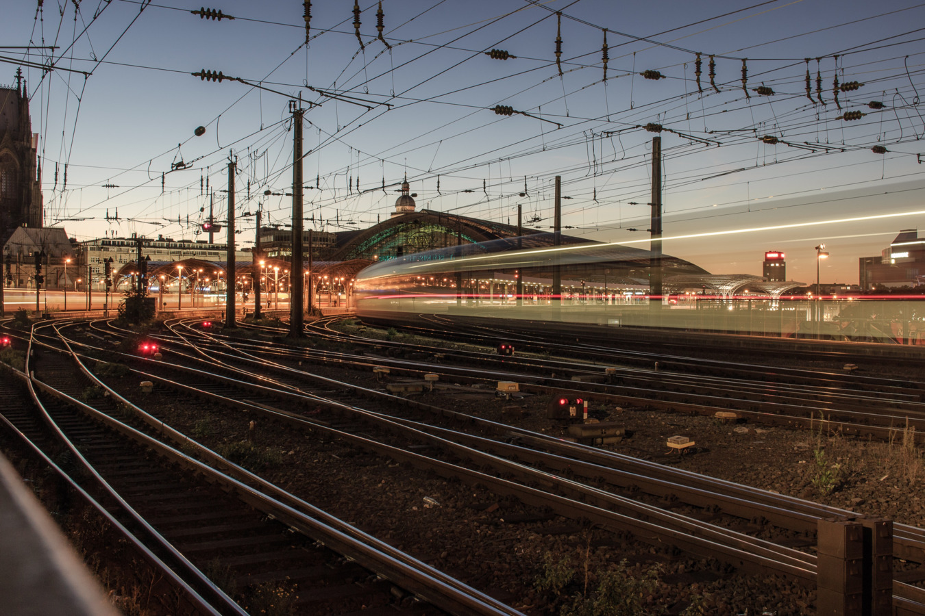 A blurred, long-exposure shot at dusk shows multiple train tracks converging towards a lit train station in the background, with overhead power lines crisscrossing the frame and light trails from a moving train.