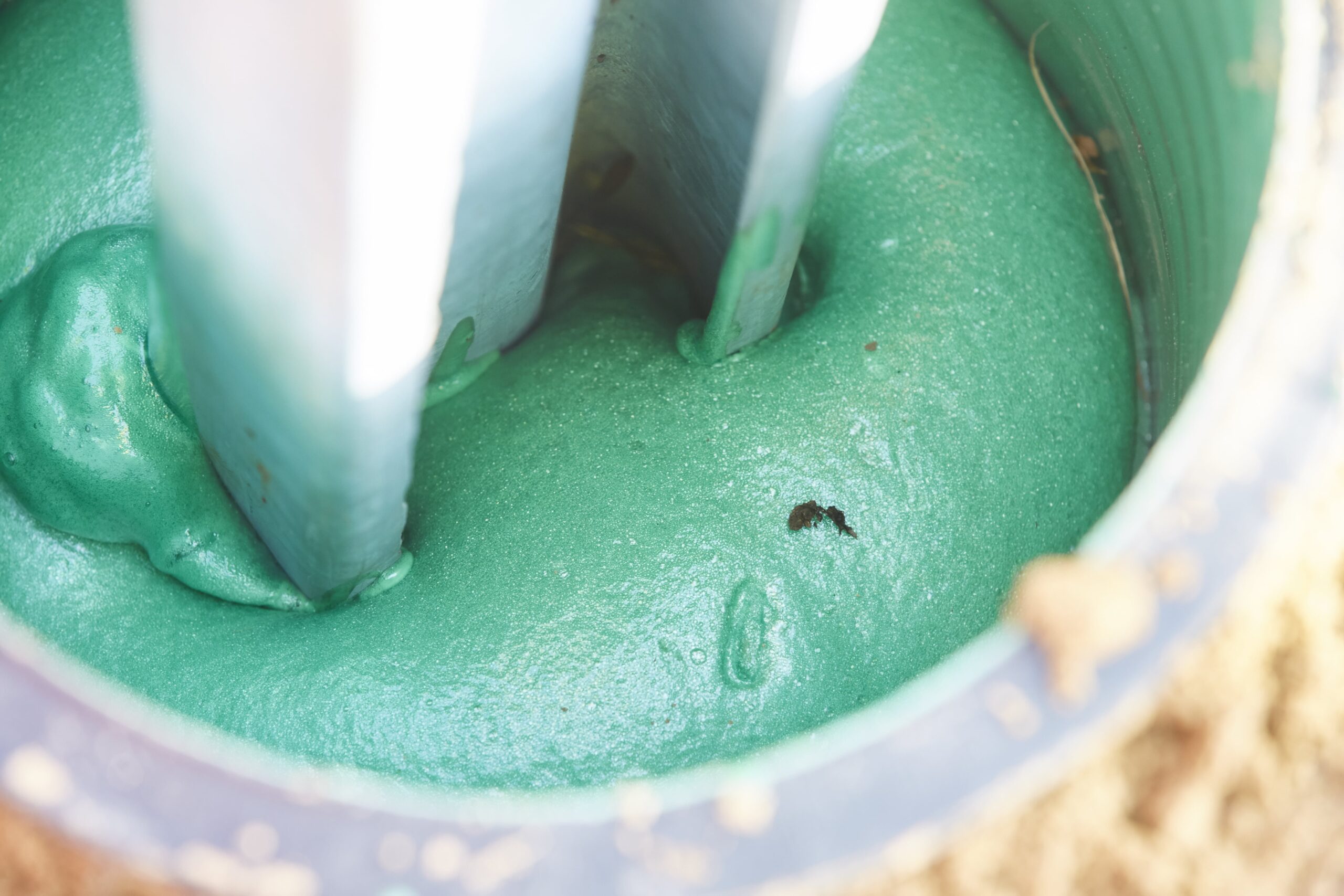 A close-up view shows a vibrant green, foamy substance being mixed in a container by two white tools, with some light brown material visible on the outer edge.