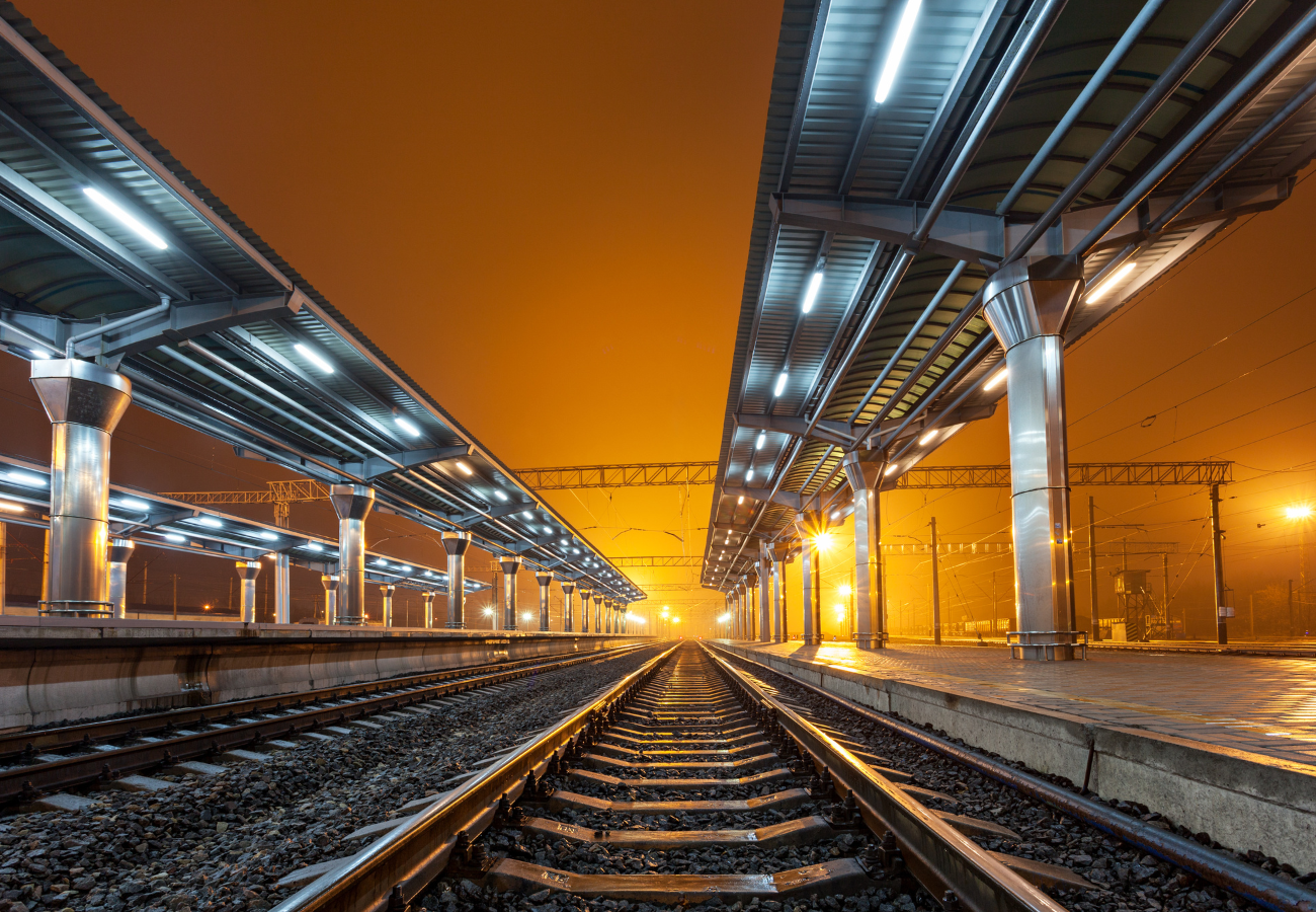 Modern railway station at night with illuminated platforms and steel canopies, viewed from ground level along the tracks.