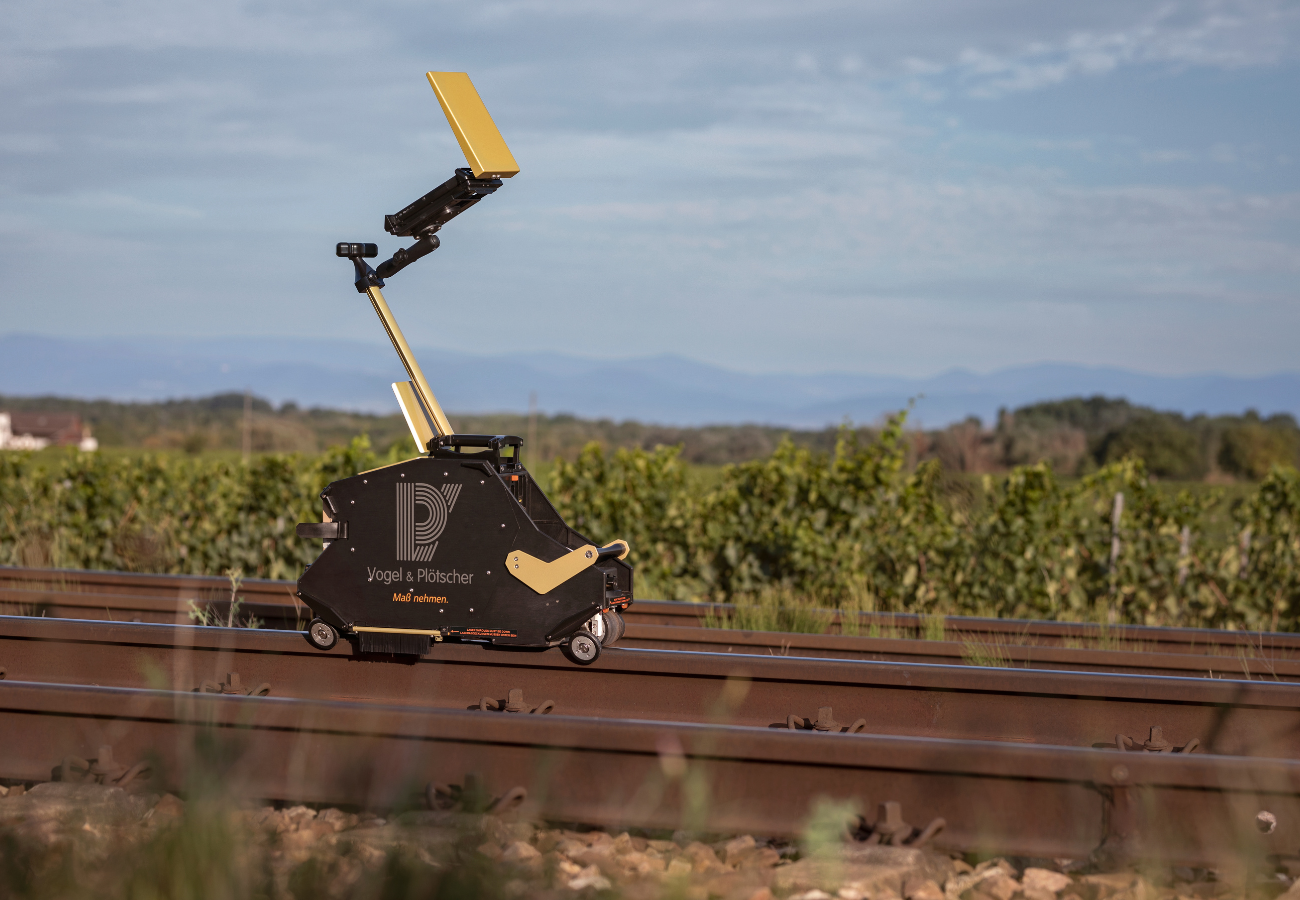 A black and gold, rail-mounted robot with an extendable arm topped with a golden panel travels on train tracks through a vineyard.