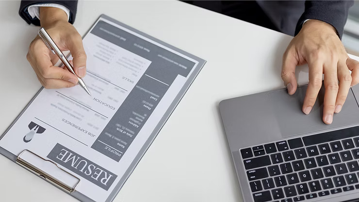 An overhead shot shows a person's hands, one holding a silver pen poised over a resume on a clipboard, and the other resting on a laptop's touchpad, all on a clean white desk.