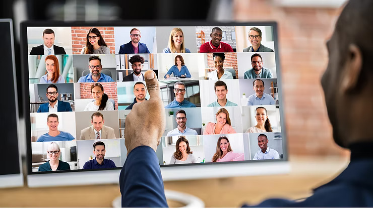 A person viewed from behind points towards a computer screen displaying a video conference call with multiple participants in a grid layout.