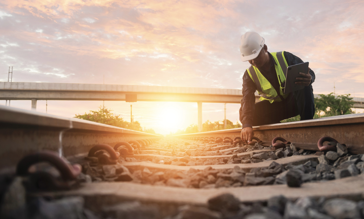 An infrastructure worker in a hard hat and safety vest inspects railroad tracks with a tablet at sunset.