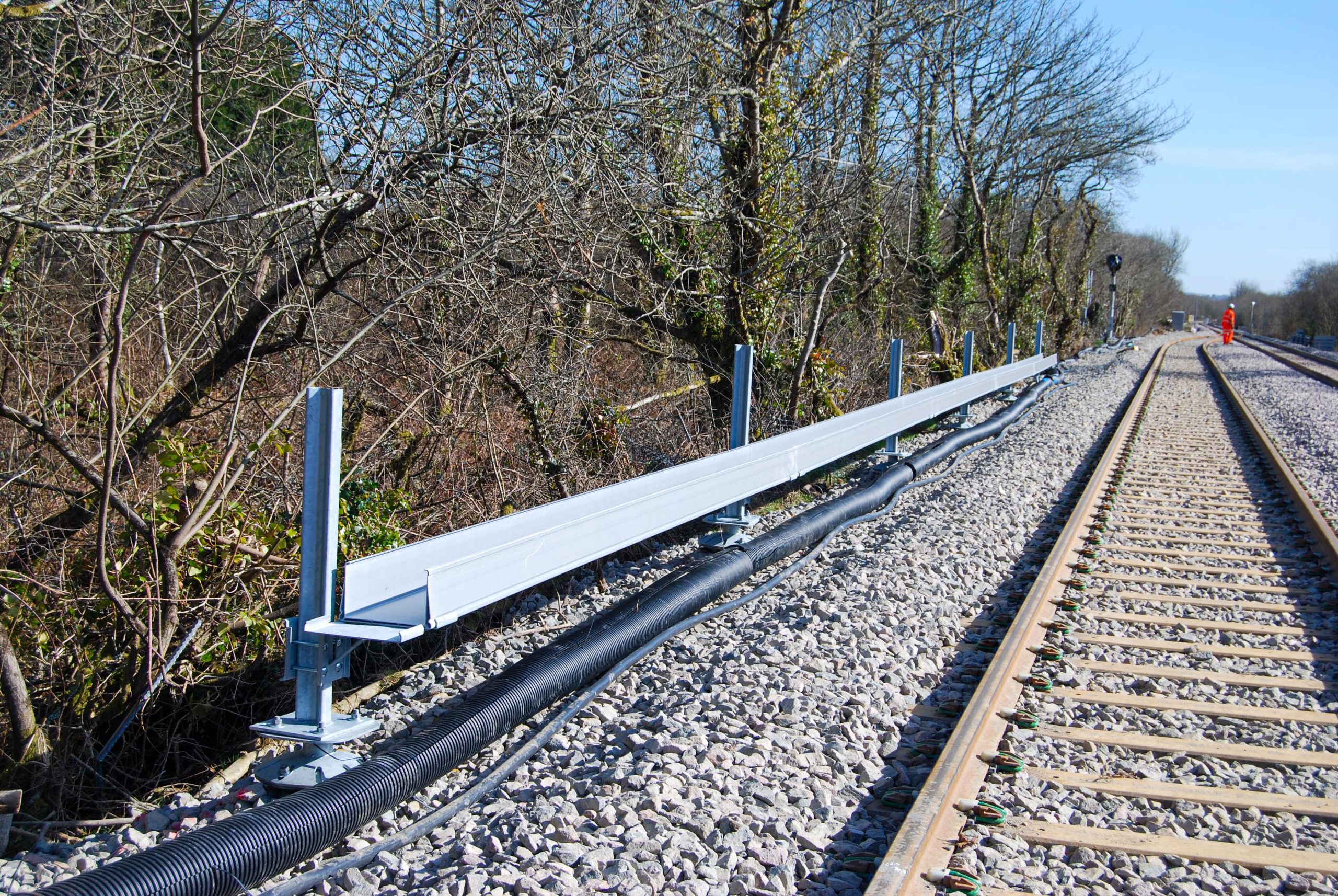 A new metal trough cable management system is installed alongside active train tracks, with bare trees bordering the railway and a person in an orange vest visible in the distance.