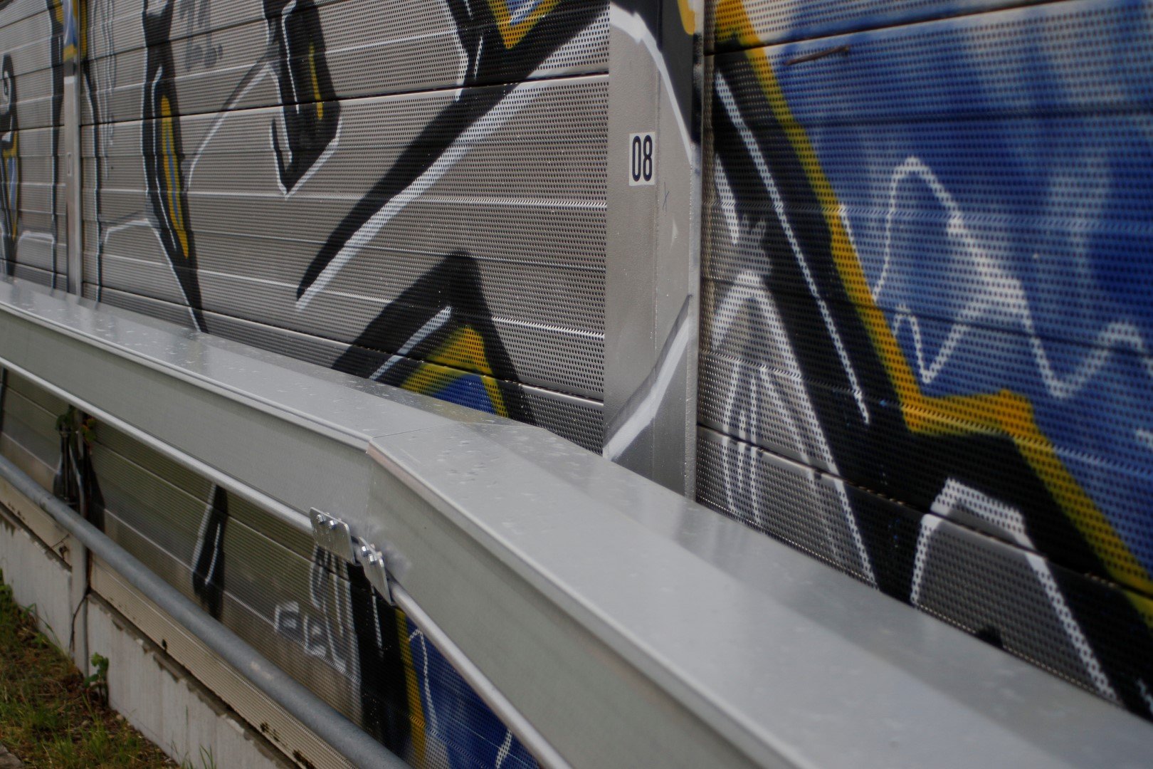 A close-up view of a metal sound barrier alongside a road, adorned with vibrant graffiti in shades of blue, black, and yellow.