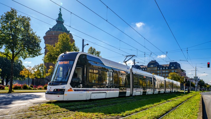 A modern, white and black tram with "Sonderfahrt" displayed on the front travels along tracks set in a grassy median under a clear blue sky. In the background, a historic water tower building and other classical architecture are visible.