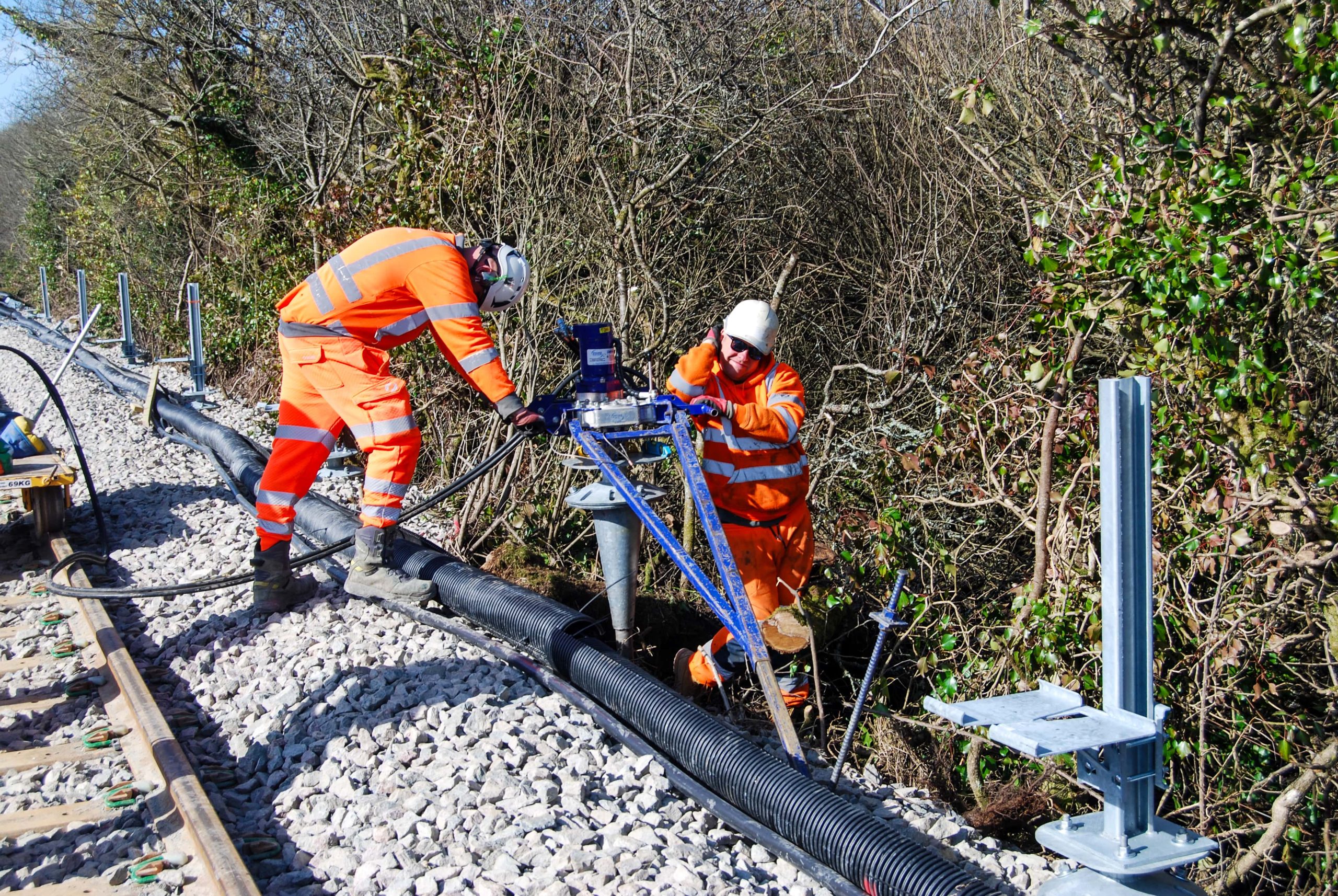 Two construction workers in orange high-visibility gear are operating machinery next to a black corrugated pipe and newly laid train tracks, surrounded by dense winter foliage under a clear sky.