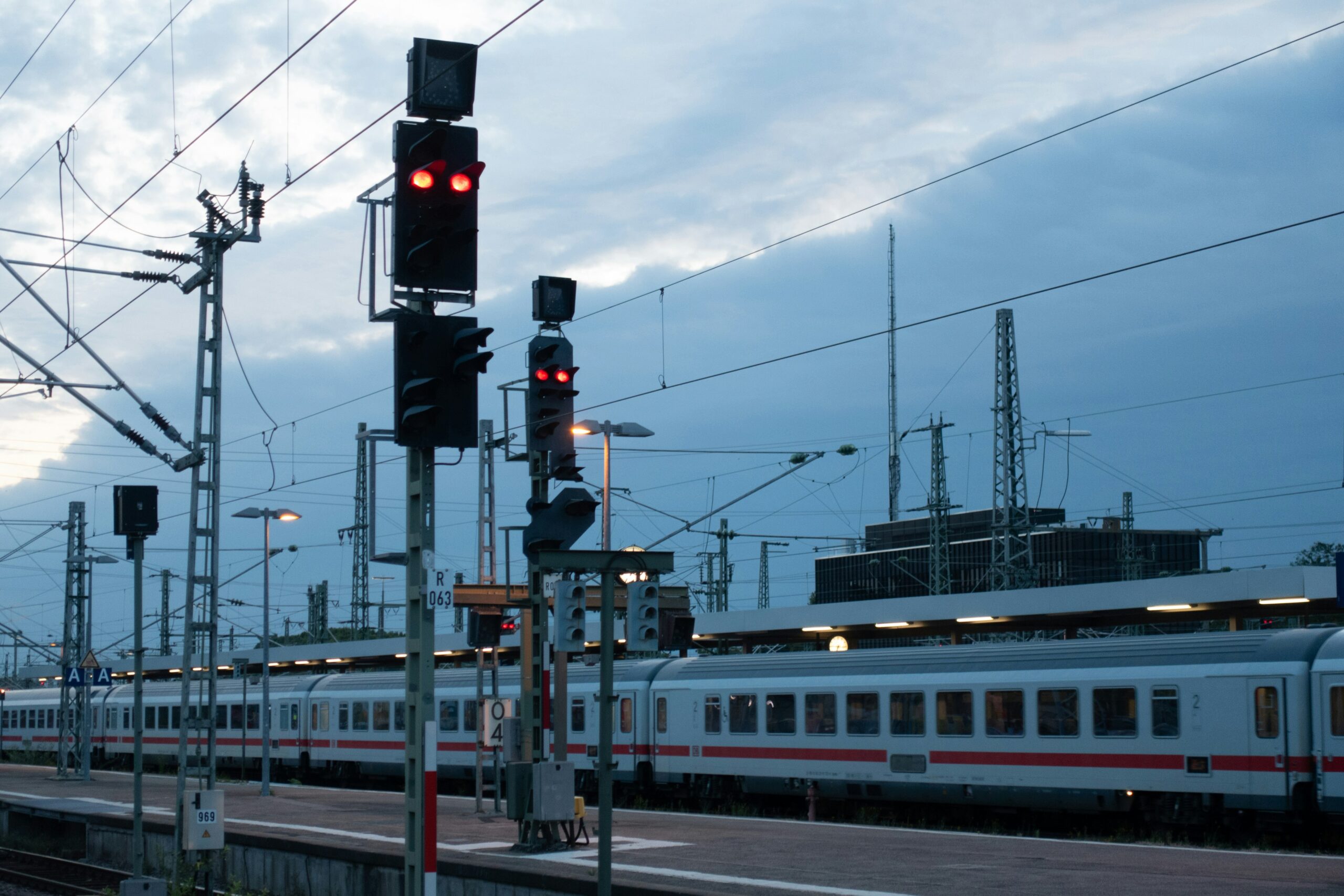 A red-light signal stands prominently on a train platform next to a white and red passenger train under an overcast sky with numerous overhead power lines and structures.