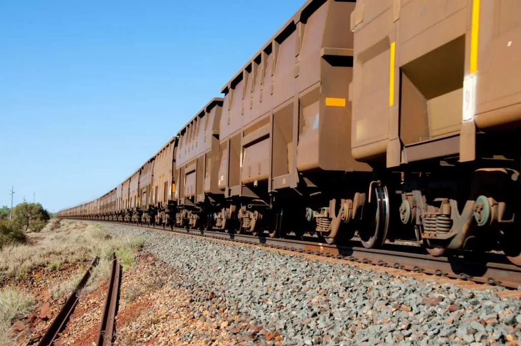 A long freight train with numerous brown railcars moves along a railway track under a clear blue sky.