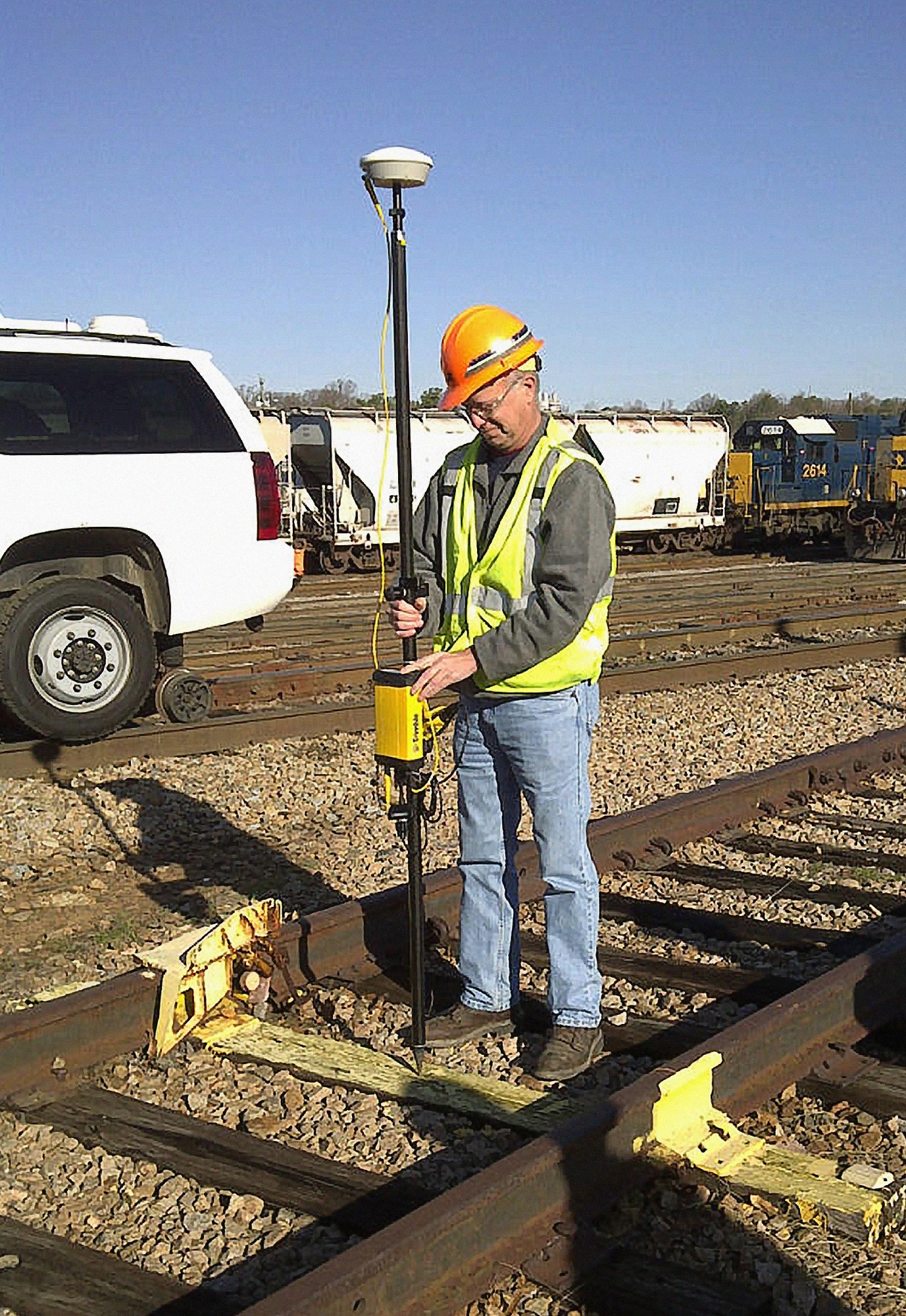 A man in an orange hard hat and safety vest operates a yellow GPS surveying pole on a set of railroad tracks, with a white SUV and train cars visible in the background under a clear sky.