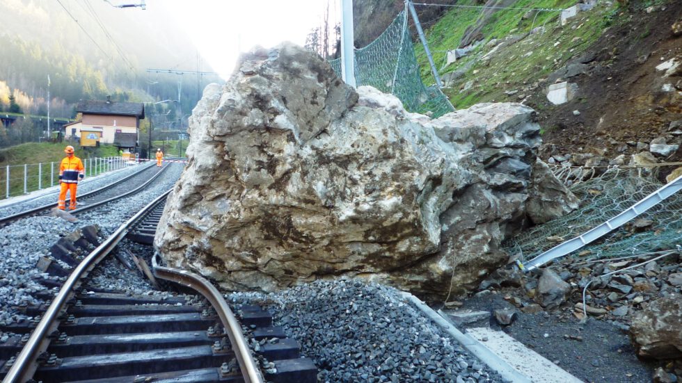 A gigantic, rough-textured boulder lies across train tracks, causing a partial derailment, while two workers in orange safety attire stand near a protective mesh barrier on an adjacent hillside under bright sunlight.