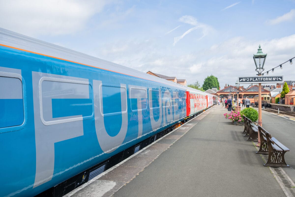 A bright blue and silver train with the word "FUTURE" emblazoned on its side is parked at a historic-looking train station platform, with people walking in the background and a sign for "Nº2 PLATFORM Nº 1".