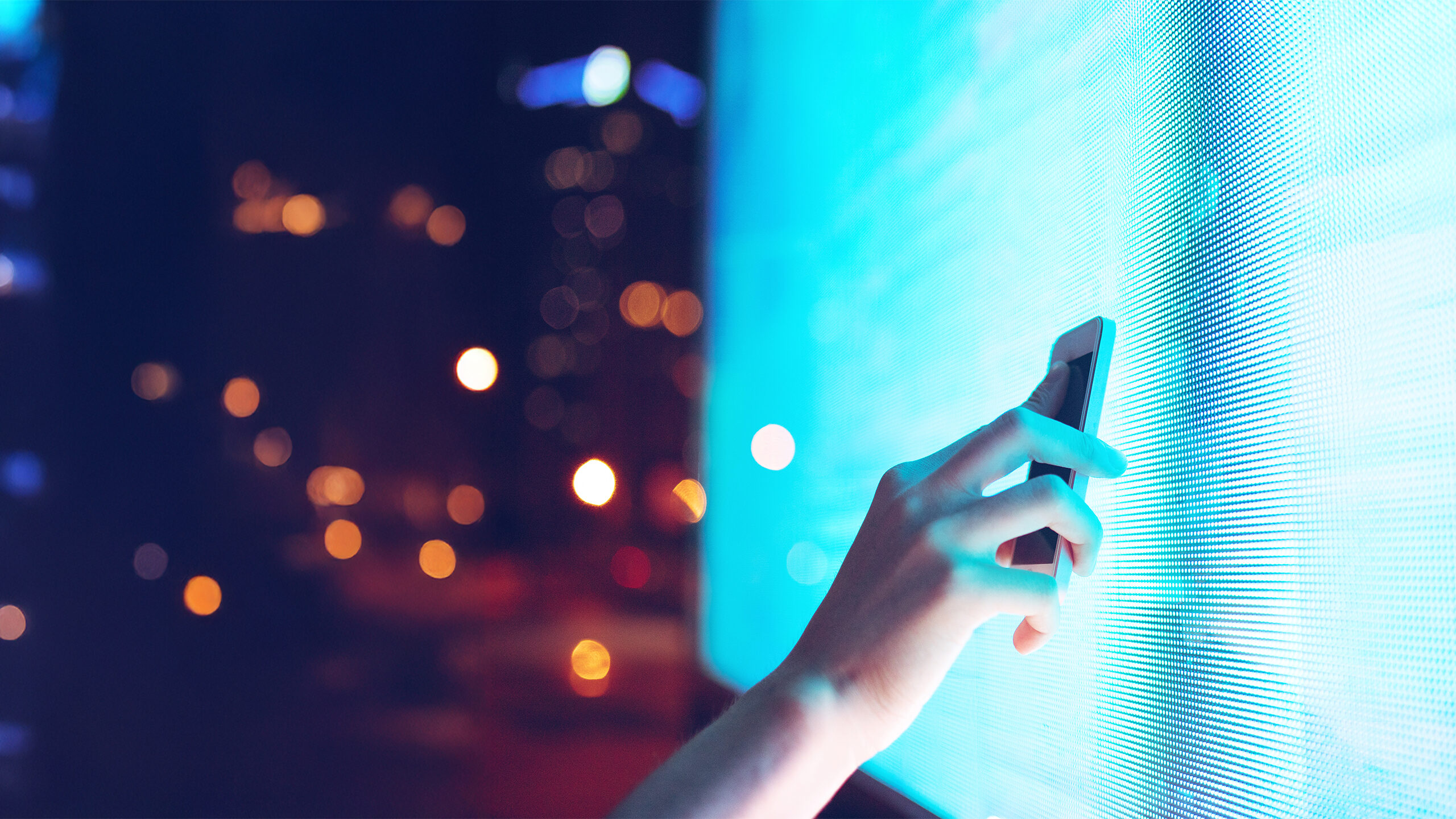 A human hand holding a smart phone up to a bright LED screen