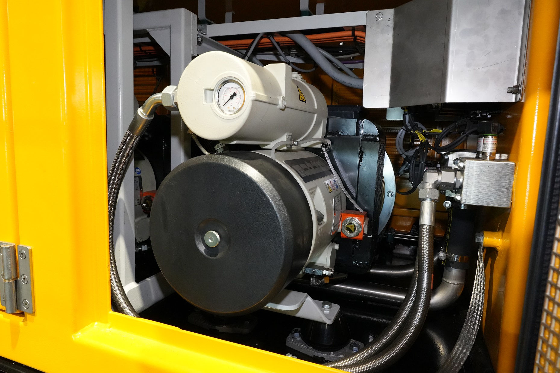 A metallic industrial component, possibly a pump or motor, is shown in close-up with a pressure gauge and hoses connected, set within a yellow structure.