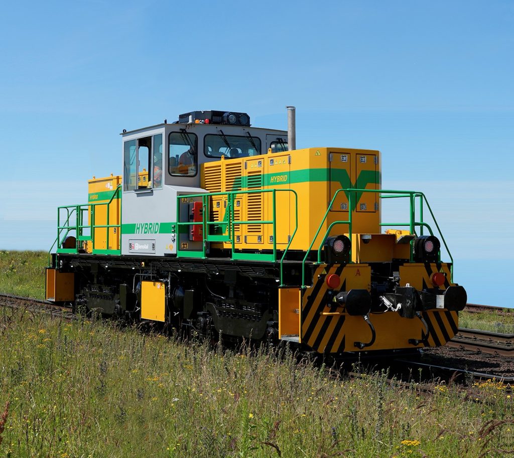 A grey, green, and yellow hybrid shunter locomotive sits on train tracks in a grassy, open field under a clear blue sky.