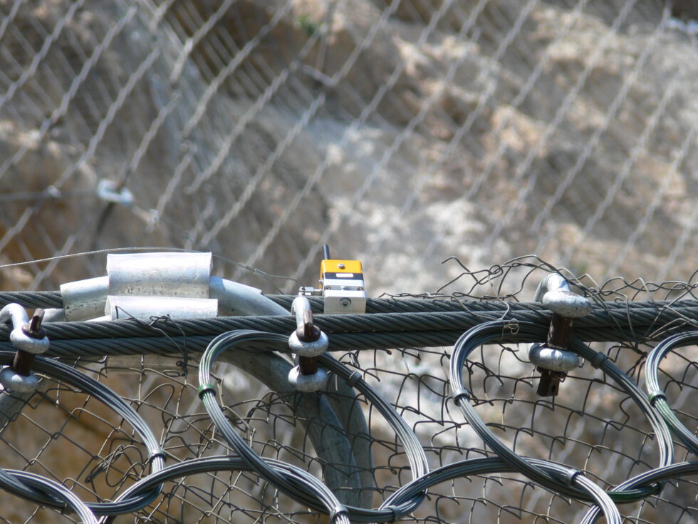 A close-up view shows a metal mesh barrier system, likely designed for rockfall protection, with a small yellow and black sensor device attached to the top cable, all against a blurred rocky background.