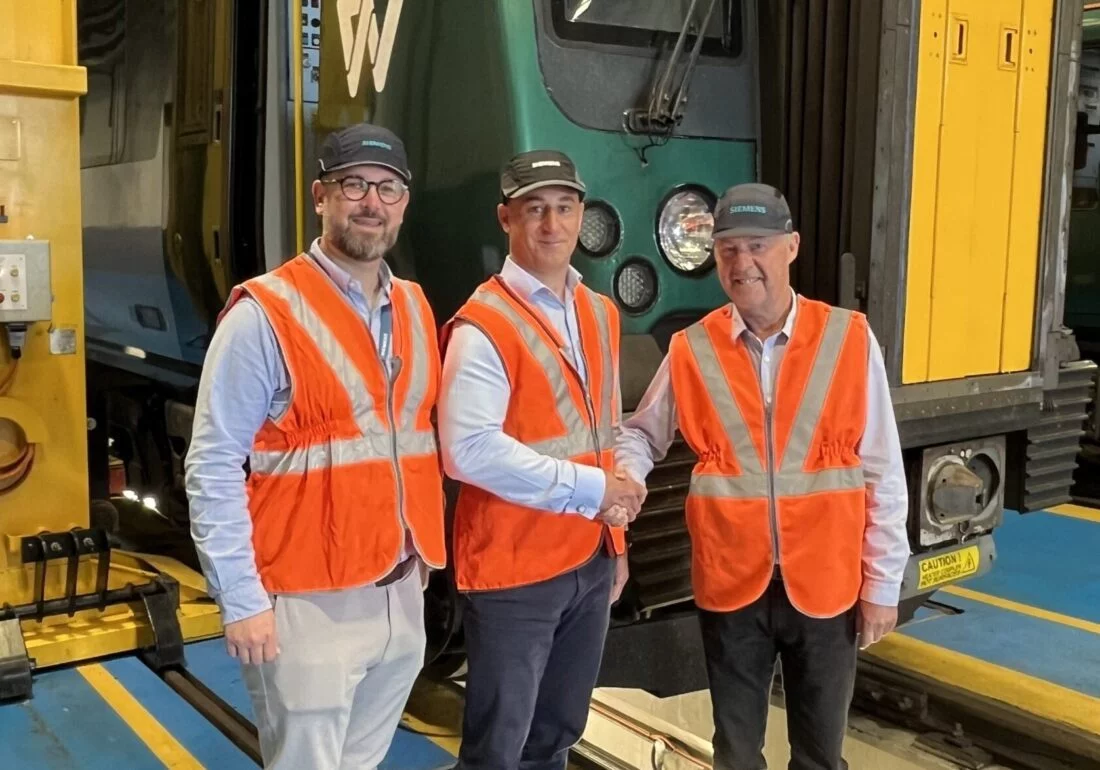 Three men in safety vests and hats shake hands in front of a train.