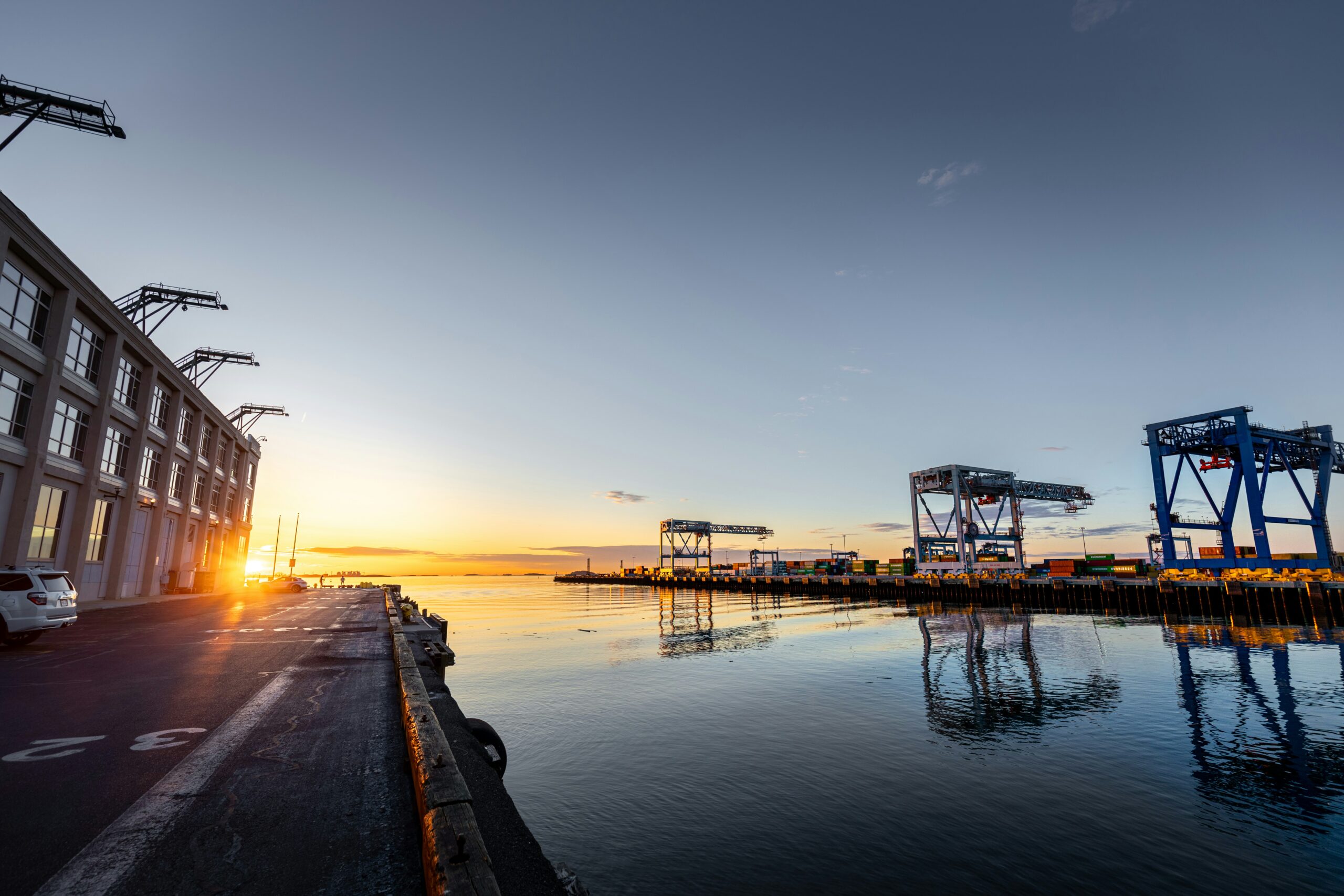 A panoramic view of a port terminal at sunset, featuring a calm body of water reflecting the sky and large blue container cranes in the distance. A multi-story building is visible on the left side, and the setting sun casts a golden glow over the water.
