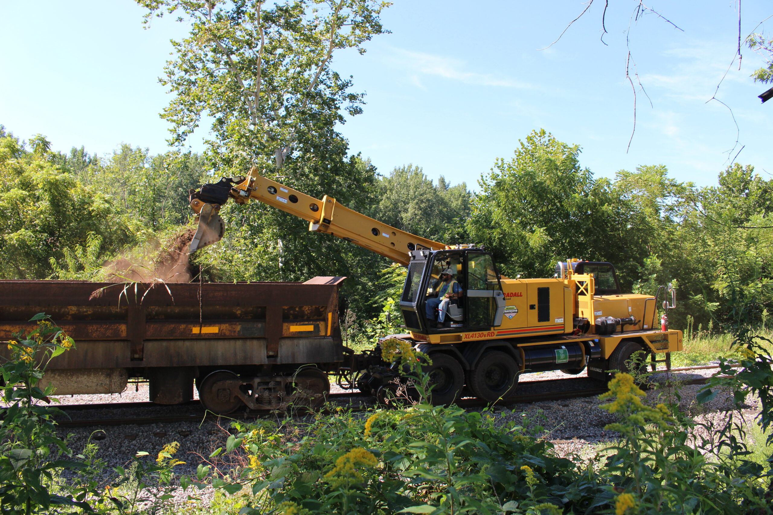 A yellow railway maintenance vehicle on tracks loads dirt into a rust-colored railcar amidst green trees and yellow flowers.