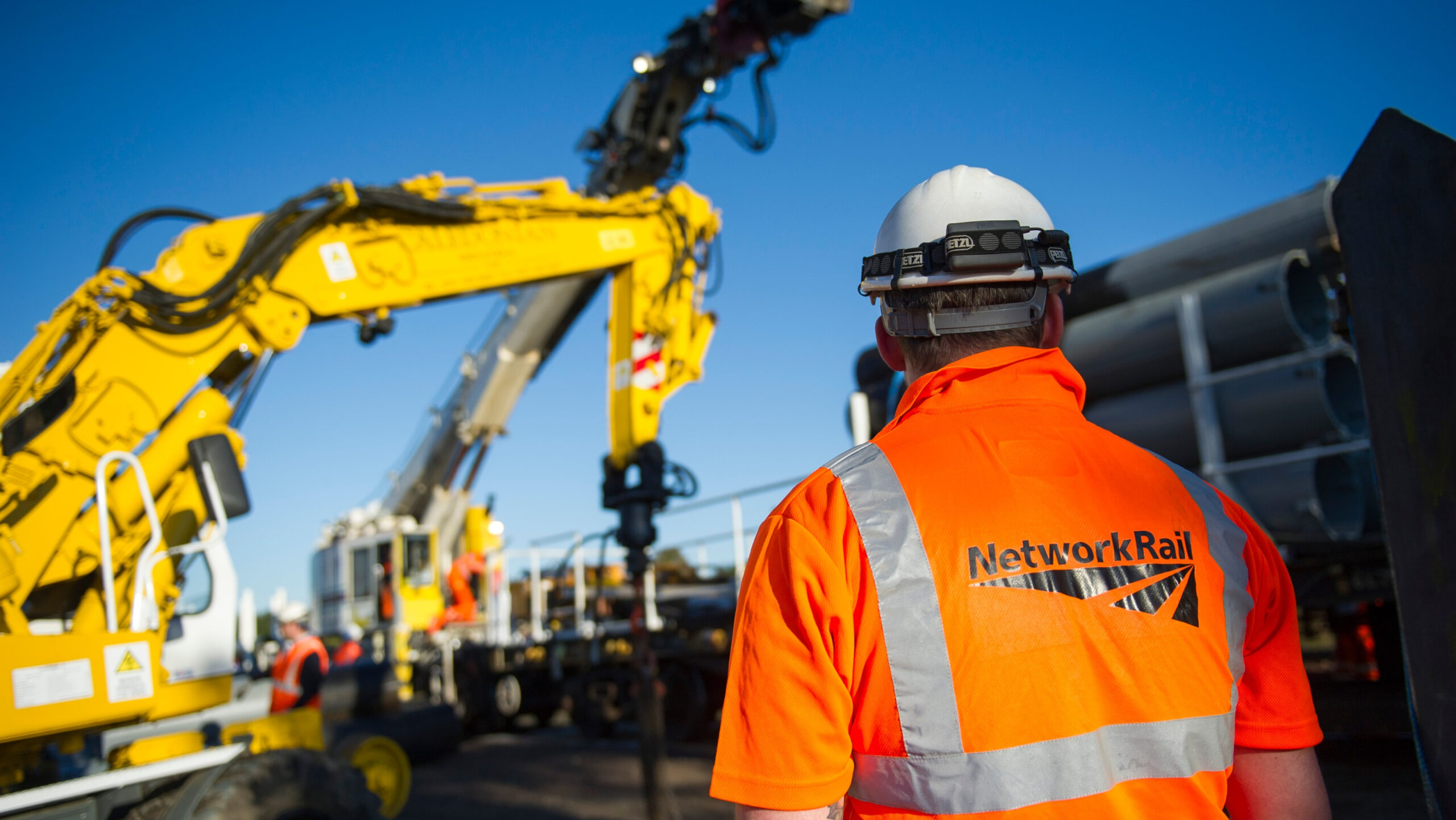 A network rail engineer in an orange hi-vis jacket looking in the direction of a yellow crane during engineering work