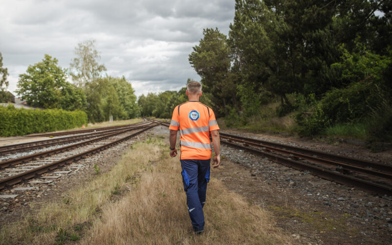 Man walking with high vis clothing on by train tracks with overcast sky.